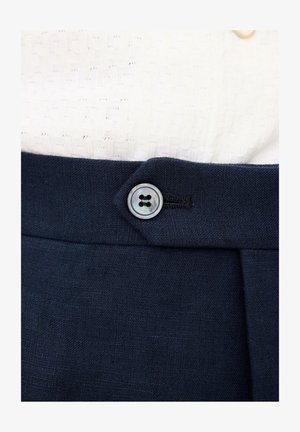 Lähikuva tummansinisten housujen vyötäröstä, jossa on kiinnitettynä yksi pyöreä helmiäishohtoinen nappi, yhdistettynä teksturoituun valkoiseen neulepaitaan.