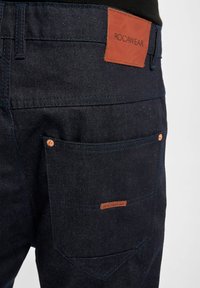 Temno modre jeans hlače iz denima z rjavim usnjenim logotipom ROCAWEAR na hrbtni strani. Vključujejo standardno zadnjo žep z bakrenimi dodatki.