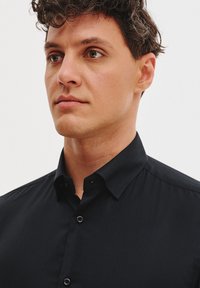 Czarna koszula na guziki z kołnierzykiem, wykonana z gładkiego materiału i o dopasowanym kroju. Dwa guziki widoczne z przodu. Gładkie tło.