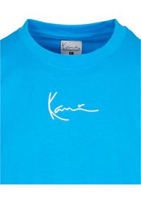 Tirkīza kokvilnas T-krekls ar apaļu kakla izgriezumu un baltu izšūtu logo "Kari" uz krūšu daļas. Gluda tekstūra, ikdienišķs dizains.