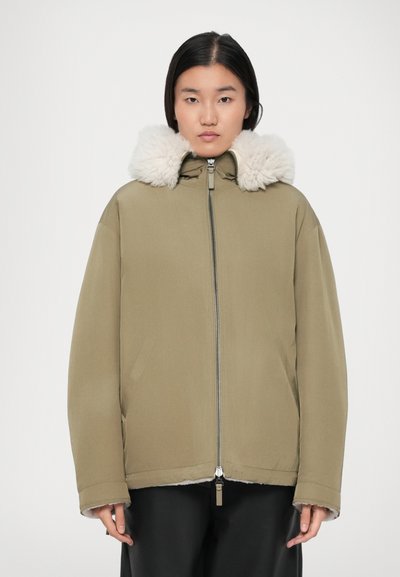 Vrouw in een olijfgroene ritsjack met een grote fluffy witte kraag en zwarte broek, staande tegen een effen achtergrond.