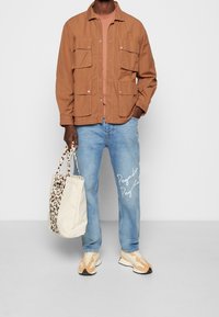 Brun lättviktig jacka med fyra fickor, rosa t-shirt, ljusblå jeans med vit text, och beige sneakers. Inkluderar en toteväska med leopardmönster.