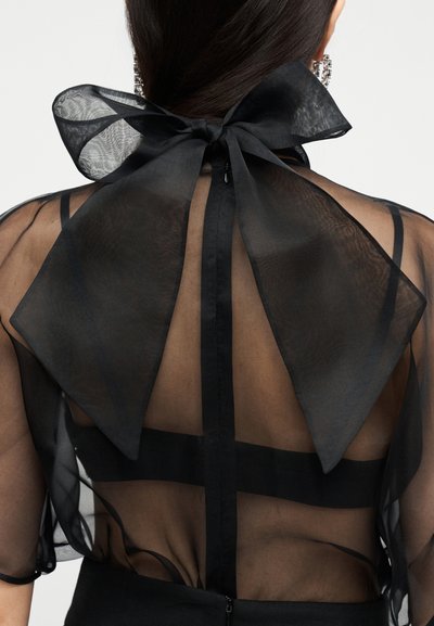 Η πλάτη γυναίκας που φοράει διάφανο μαύρο τοπ με μεγάλο μαύρο φιόγκο δεμένο στον λαιμό και ορατές μαύρες τιράντες σουτιέν από κάτω.