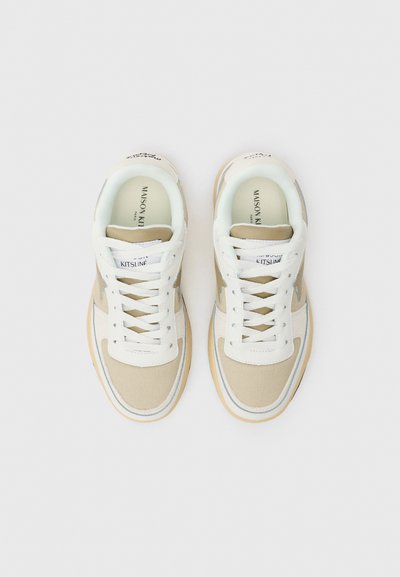 Beige ja valged tennisetossud, millel on riidest pealispind, valged paelad ja kerge tallaga. Tootemärk keelel ja sile tekstuur.