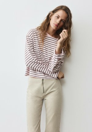 Jeune femme aux longs cheveux ondulés portant un haut rayé rose et noir à manches longues et un pantalon beige, debout contre un mur blanc uni.