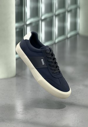 Marineblaue BOSS-Sneaker mit weißer Sohle und Fersendetail, schwarzen Schnürsenkeln, schwebend vor einem Hintergrund aus grauem Boden und Glaswand.