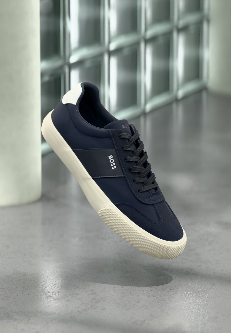 Baskets BOSS bleu marine avec semelle blanche et détail au talon, lacets noirs, en suspension dans les airs sur fond de sol gris et mur en verre.