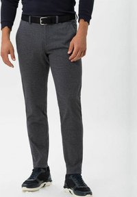 Man wearing slim-fit gray patterned trousers, black belt, black sneakers, and a dark long-sleeve top, standing against a plain background.