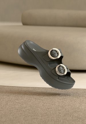 Sandale plateforme noire à enfiler avec deux grandes boucles rondes, semelle intérieure texturée, présentée flottante au-dessus d'un tapis beige et d'un canapé en arrière-plan.