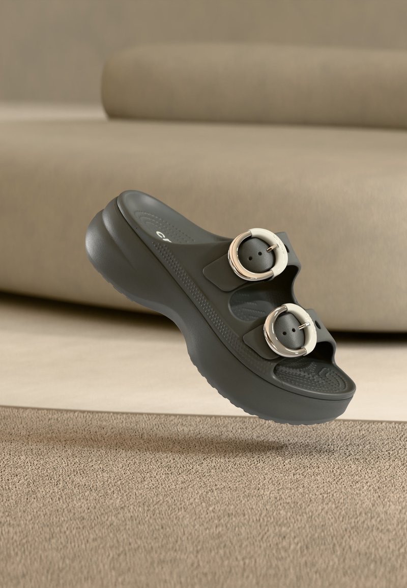 Μαύρο πλατφόρμα σάνταλ με δύο μεγάλες στρογγυλές αγκράφες, υφή στη σόλα, εμφανίζεται να αιωρείται πάνω από μπεζ χαλί και φόντο καναπέ.