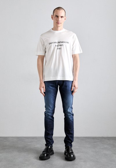 Weißes Baumwoll-T-Shirt mit schwarzem Druck "EMPORIO ARMANI MILANO 1981", kombiniert mit dunkelblauen Slim-Fit-Jeans und schwarzen klobigen Schuhen.