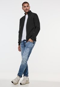 Černá zipová bunda s texturovaným předním dílem, nošená přes bílé tričko, spárovaná se světle modrými džíny a světlými teniskami.