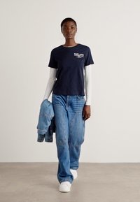 T-shirt blu navy a maniche corte con logo bianco, sovrapposto a un top bianco a maniche lunghe. Abbinato a jeans in denim azzurri a vestibilità ampia e sneakers bianche.