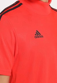 Punainen Adidas urheilupaita, jossa musta logo rinnassa ja mustat kolme raitaa olkapäällä, henkilö pukeutunut siihen.