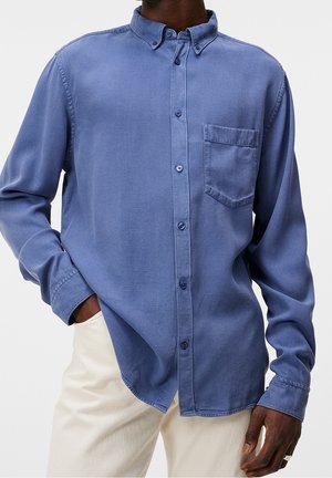 Homme portant une chemise bleue à manches longues avec une poche poitrine et un pantalon crème, main dans la poche et coupe décontractée.