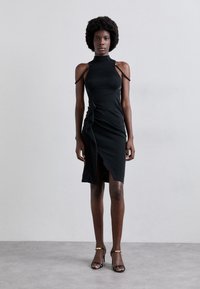 Vestido negro ajustado de cuerpo con cuello alto y hombros descubiertos. Presenta un detalle retorcido en la cintura y una abertura alta hasta el muslo. Textura suave.