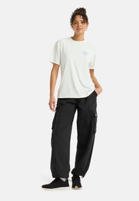 Camiseta ligera de crema con logotipo azul, combinada con pantalones cargo negros holgados. El atuendo cuenta con múltiples bolsillos y puños elásticos.