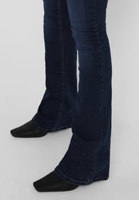 Dark blue flared jeans with a slight sheen, featuring a fitted waist and subtly frayed hem. Paired with pointed black shoes.