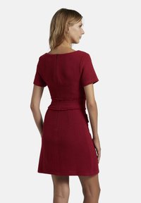 Rotes, strukturiertes Kleid mit kurzen Ärmeln, taillierter Taille und geradem Rock. Verfügt über einen Reißverschluss auf der Rückseite und fransige Saumakzente.