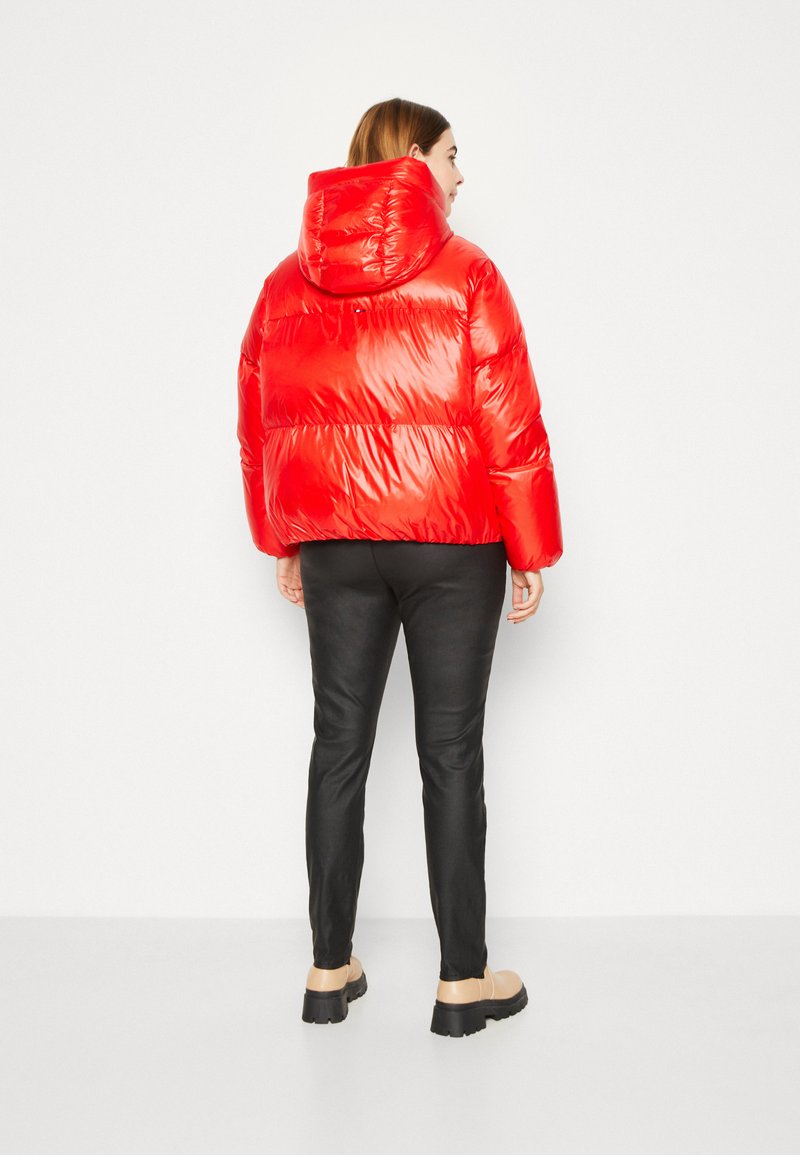 Giacca a vento rossa con una texture lucida e cappuccio, caratterizzata da una trapuntatura orizzontale. Indossata con pantaloni neri e scarpe beige. Vista posteriore.