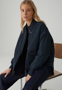 Blonde Frau, die auf einem Stuhl sitzt, trägt eine dunkelblaue Bomberjacke, ein schwarzes Oberteil und eine dunkle Jeans vor einem neutralen Hintergrund.