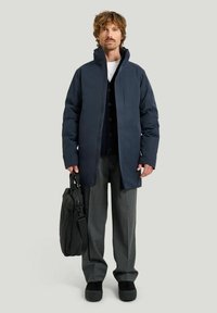 Marinblå puffjacka med hög krage, grå greppbyxor och svarta skor, hållande en svart väska. Har knappad innerskal och dragkedja.