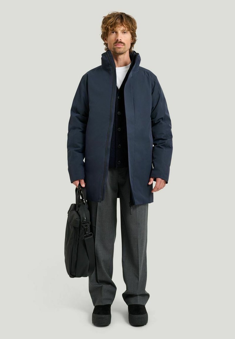 Marinblå puffjacka med hög krage, grå greppbyxor och svarta skor, hållande en svart väska. Har knappad innerskal och dragkedja.