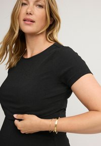 Top nero a maniche corte con tessuto texturizzato, scollatura a girocollo e design aderente. Un braccialetto dorato è indossato al polso.
