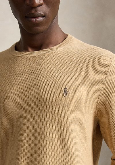 Primo piano di una persona che indossa un maglione beige lavorato a maglia con un piccolo logo ricamato con cavallo e fantino sul petto.