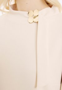 Cremfarbene Bluse mit hohem Kragen, der an der Halslinie mit einem goldenen, ovalen Akzent verziert ist, und einem weichen, strukturierten Stoff. Elegantes Design mit einem Bindedetail.