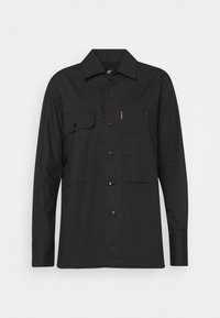 Camicia utilitaria nera con maniche lunghe, chiusura a pulsante, due tasche sul petto e cuciture di accento giallo sulla tasca sinistra. Tessuto liscio.