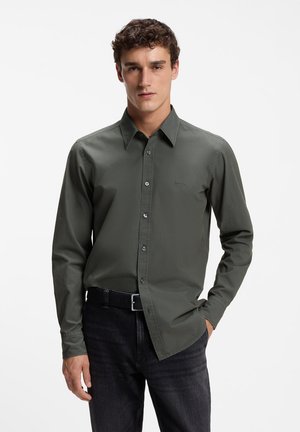Junger Mann, der ein olivgrünes Hemd mit Knöpfen und dunkle Jeans mit einem schwarzen Gürtel trägt, steht mit einer Hand in der Tasche.