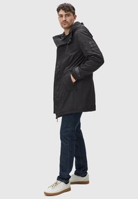 Svart parkasjacka med huva, med dragkedja fram, sidofickor och strukturerade ärmlappar. Matchas med blå jeans och vita sneakers.