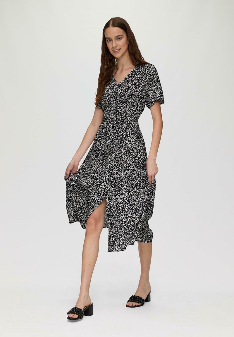 Robe noire à fleurs avec des manches courtes, décolleté en V et fente haute jusqu'à la cuisse, associée à des sandales à talons carrés noires. Texture du tissu lisse.
