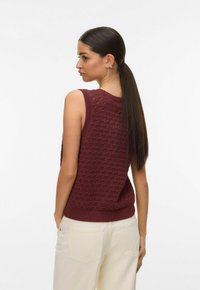 Femme avec une longue queue de cheval noire portant un haut en maille bordeaux sans manches et un pantalon clair, regardant par-dessus son épaule vers la gauche.