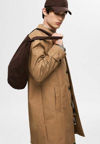 Cappotto impermeabile beige con chiusura a bottoni, tasche laterali e maniche arrotolate, abbinato a una borsa marrone scuro e un cappello coordinato.