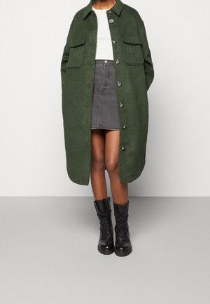 Person trägt einen langen grünen, geknöpften Mantel über einem weißen Oberteil, grauem Jeansrock und schwarzen Schnürstiefeln und steht vor einem einfarbigen Hintergrund.