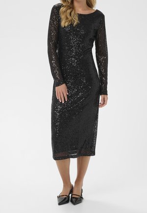 Donna che indossa un lungo vestito nero con paillettes, maniche lunghe trasparenti e décolleté nere con punta a punta e cinturini.
