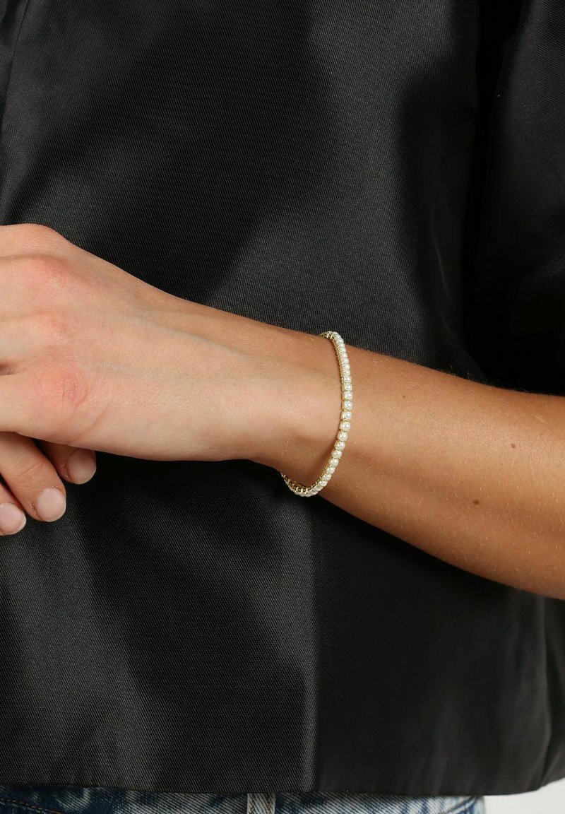 Gold-tone bracelet with small bead accents, smooth texture, slender design, resting on a wrist against a dark fabric background.