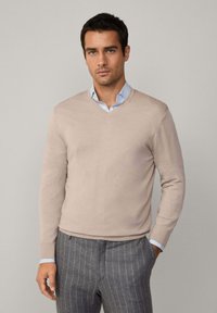 Béžový sveter s výstrihom do V vyrobený z ľahkého materiálu, s dlhými rukávmi a rebrovanými manžetami, v kombinácii so šedými nohavicami s prúžkom a svetlomodrou košeľou.