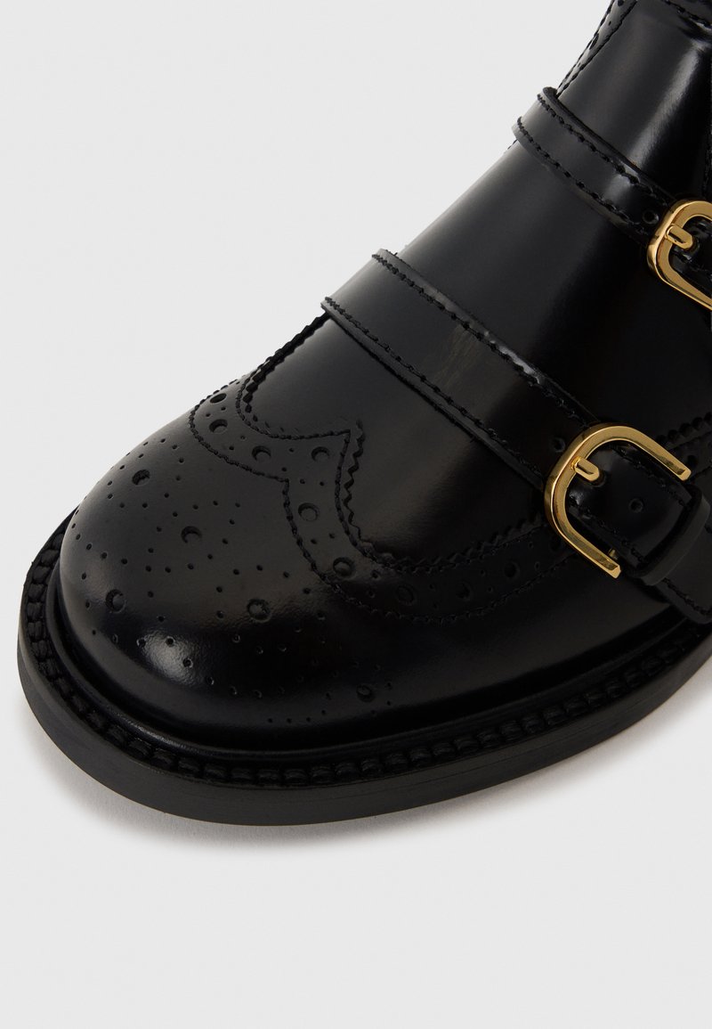 Scarpa in pelle nera con fibbie, dettagli perforati e finitura liscia. Presenta due fibbie color oro e una punta arrotondata.