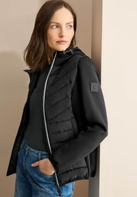 Giacca nera isolante con fronte testurizzato, chiusura con zip e cappuccio regolabile. Abbinata a un top nero e jeans blu. Dettagli di design semplici.
