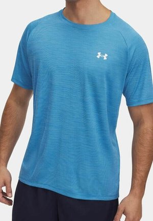Mężczyzna ubrany w jasnoniebieską sportową koszulkę z krótkim rękawem z białym logo Under Armour na lewej piersi.