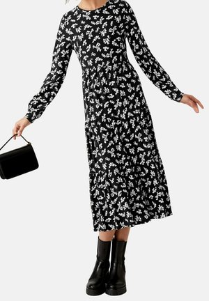 Femme portant une robe midi noire à manches longues avec un imprimé floral blanc, des bottes noires et tenant un petit sac à main noir.
