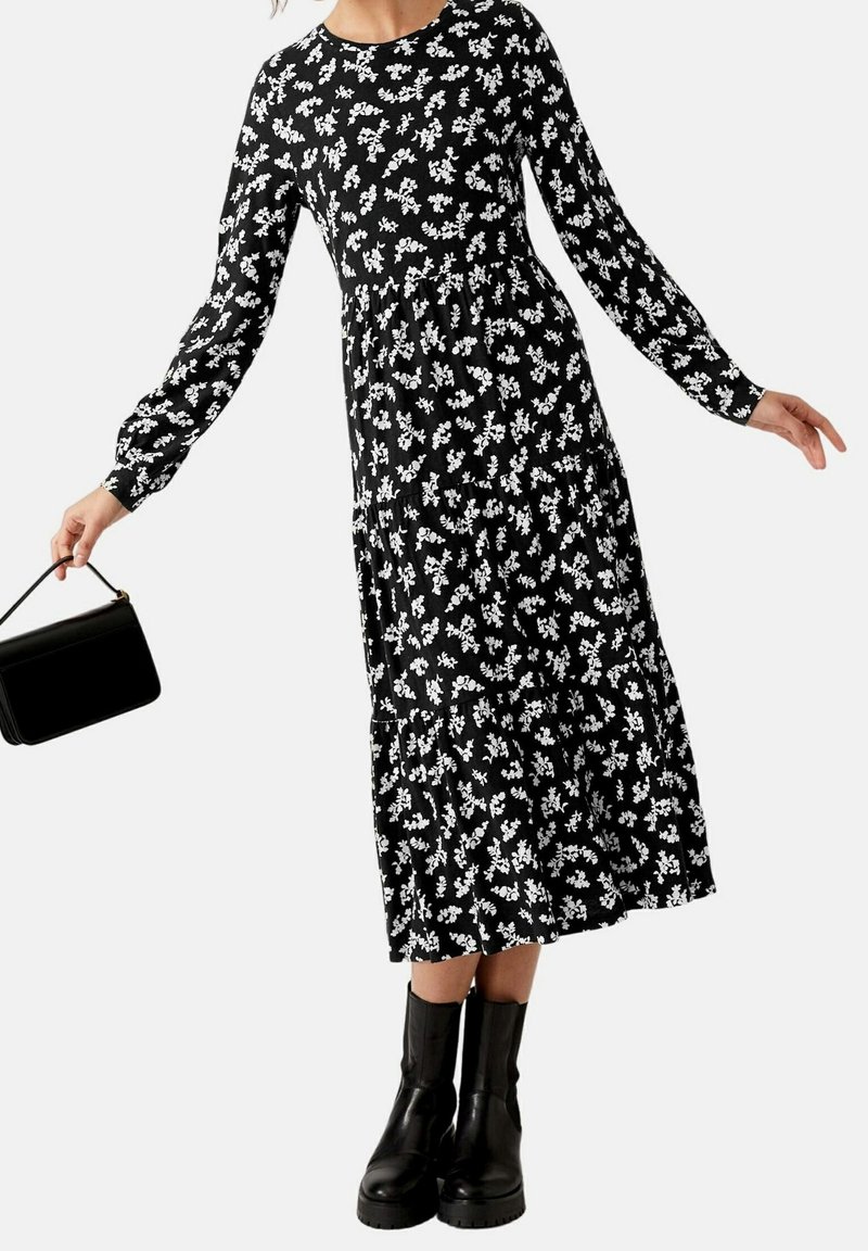 Femme portant une robe midi noire à manches longues avec un imprimé floral blanc, des bottes noires et tenant un petit sac à main noir.