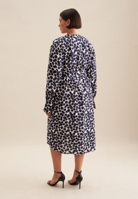 Abito a pois blu navy e bianchi con maniche lunghe, vita cintrata e lunghezza media. Tessuto liscio con una vestibilità su misura. Sandali neri con tacco alto.