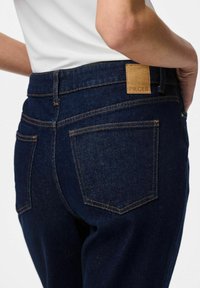 Tumši zili džinsi ar pielāgotu dizainu, ar oranžiem šuvumiem, divām aizmugurējām kabatām un brūnu ādas etiķeti ar uzrakstu "PIECES".