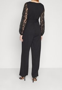 Svart jumpsuit med långa spetsärmar, figurnära midja, vida ben och dragkedja bak. Har en mjuk textur med spetsmönster.