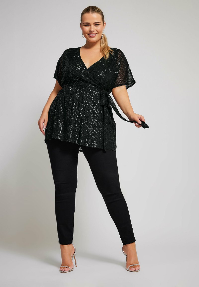 Top avvolgente nero con paillettes, scollatura a V, maniche corte e cintura regolabile, abbinato a pantaloni neri aderenti e tacchi trasparenti con lacci.