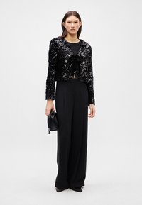 Μαύρο κολλητό σακάκι με πούλιες και ζώνη μπροστά, φορεμένο πάνω από μαύρο μπλουζάκι και φαρδιά παντελόνια, συνδυασμένο με μια μικρή μαύρη τσάντα και γόβες.